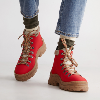 A person dressed in red hiking boots with beige accents, rope laces, green socks, and blue jeans, standing against a white background