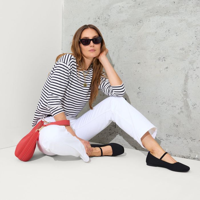 A woman in a striped shirt and white pants, wearing sunglasses and black shoes, seated with a red bag by her side