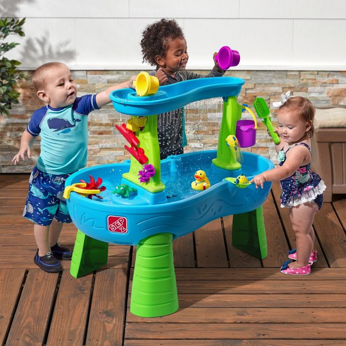 Three toddlers play with a colorful water activity table, pouring water and engaging with floating toys. The scene is set outdoors on a wooden deck.