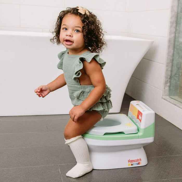 A young child, dressed in a green ruffled outfit and knee-high socks, sitting on a children's potty labeled 'Hopscotch.' The background features a bathtub and a tiled floor.