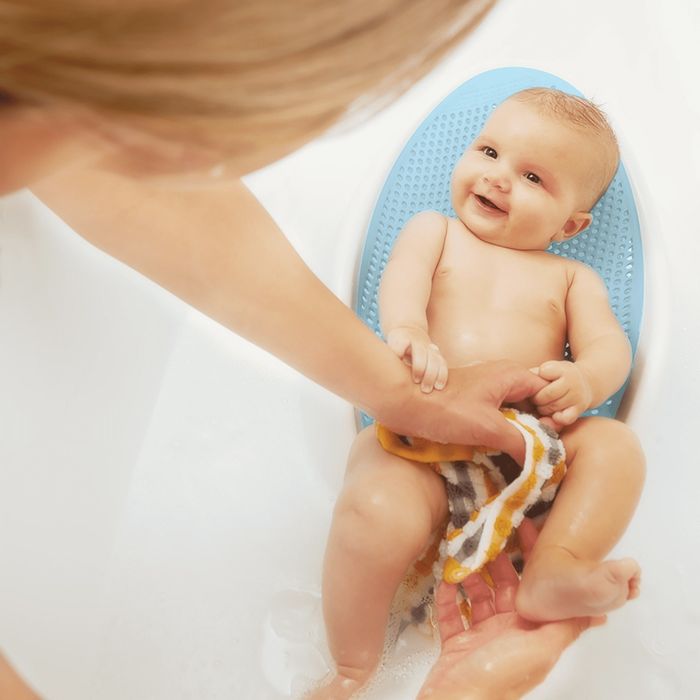 A baby smiles while being bathed in a bathtub. An adult gently washes the baby with a cloth.