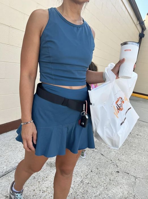 A person in a sleeveless top and skirt, holding a coffee cup and a bag, with a child in the background.