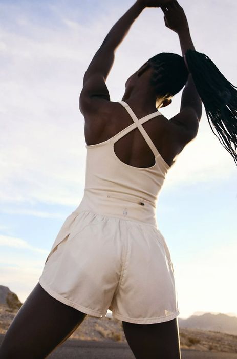 A person in white sportswear with a crisscross back, standing outdoors with their arms raised and facing away.