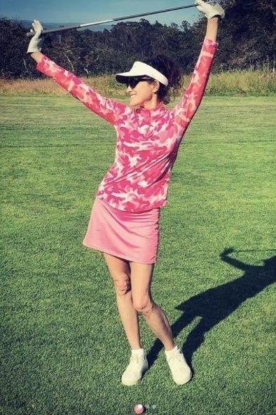 A reviewer wearing a pink patterned top and skirt celebrates a successful golf swing on the course.