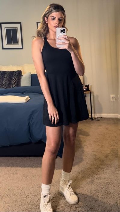 A reviewer wearing a sleeveless black dress and white sneakers, taking a mirror selfie.