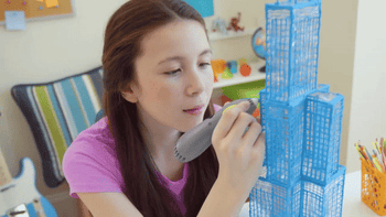 A young girl uses a 3D printing pen to build a cityscape model on a table, demonstrating a fun and creative toy for children.