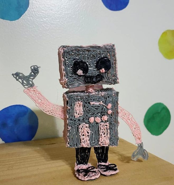 A 3D pen sculpture of a small robot with a square head, waving cheerfully on a wooden surface, set against a colorful polka dot wall in the background.