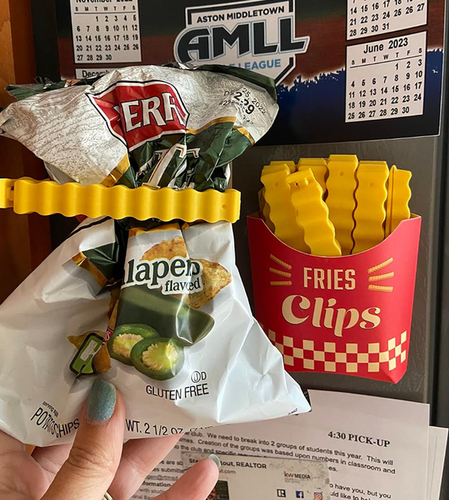 a person holding a bag of chips sealed with a plastic french fry clip, next to a magnetized holder filled with other clips