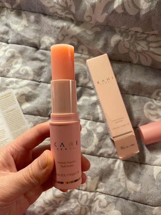 A hand holding a Kahi Wrinkle Bounce Multi Balm stick, with its packaging visible in the background