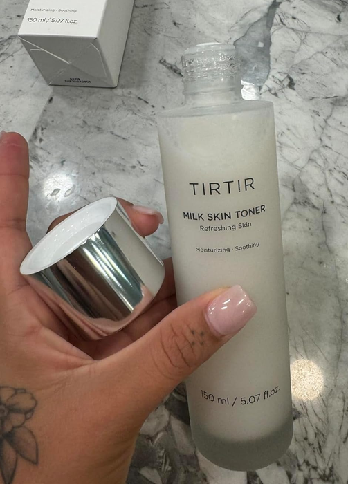 A close-up of a hand holding the TIRTIR Milk Skin Toner bottle with the lid removed, placed on a marble surface to highlight the product