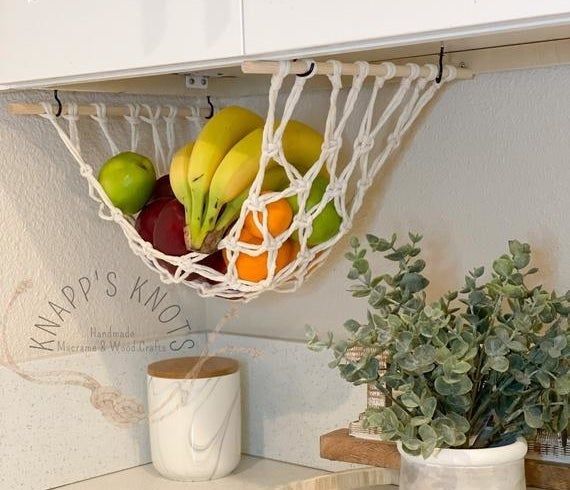 a macramé hammock suspended beneath a kitchen cabinet, filled with fresh fruits