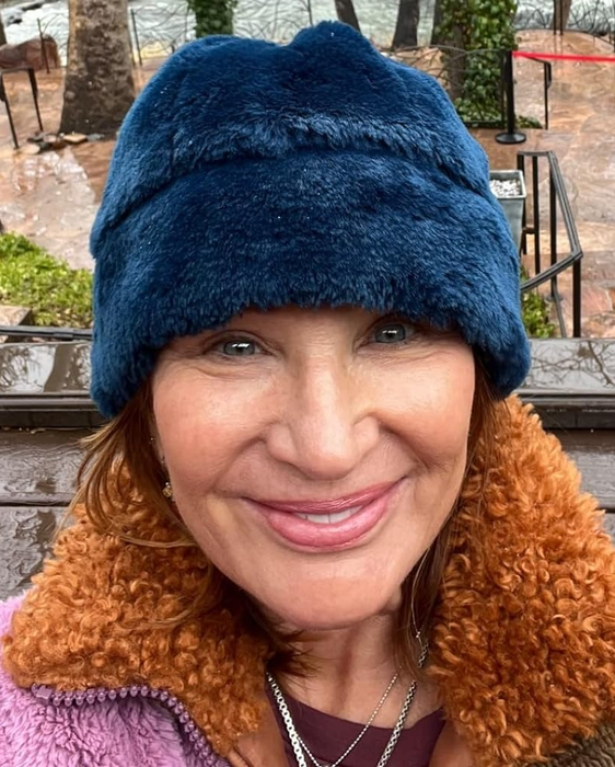 A woman in a blue hat and brown jacket smiles for a selfie with a scenic river and trees behind her