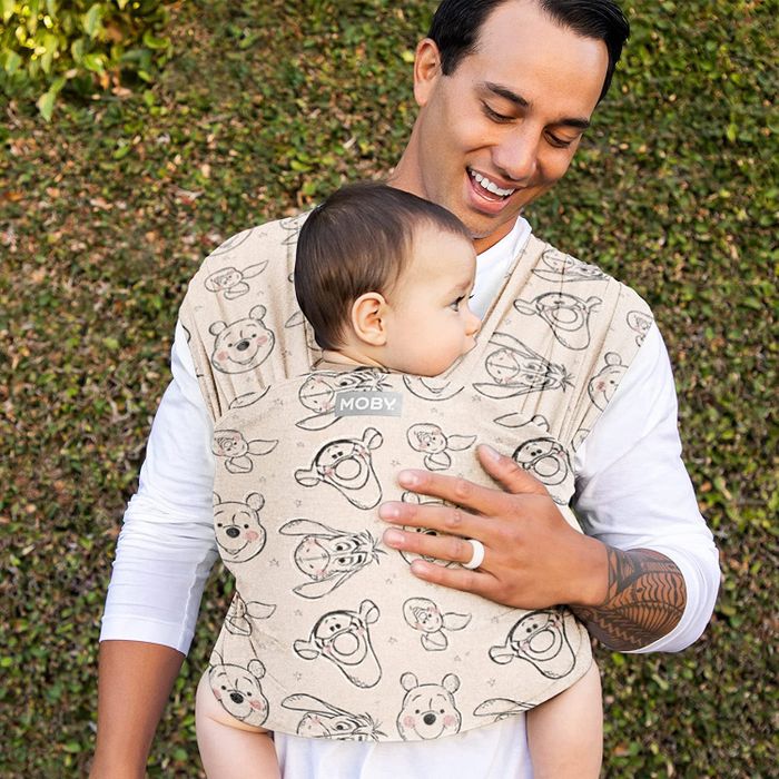 A parent wearing a tan Moby Wrap featuring Winnie the Pooh characters, keeping their baby snug and nearby.