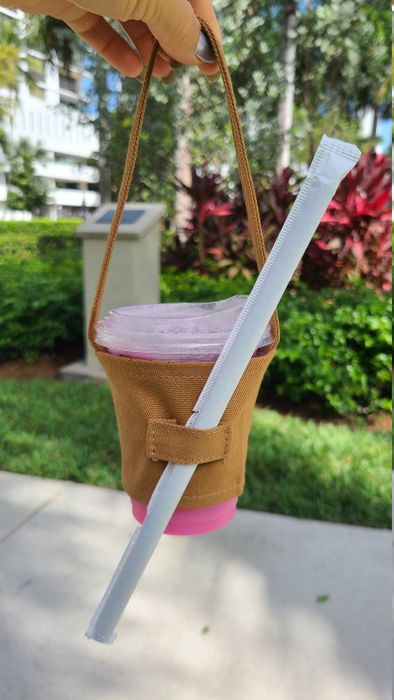 A reusable cup holder carrying a drink with a straw and lid, pictured outdoors on a bright, sunny day