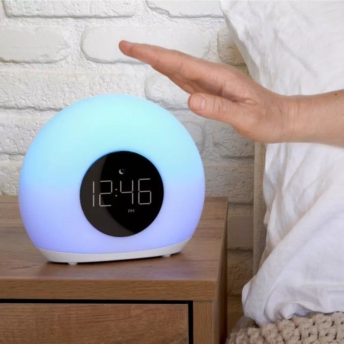 A person's hand reaching to press an alarm clock on a bedside table showing 12:46 PM