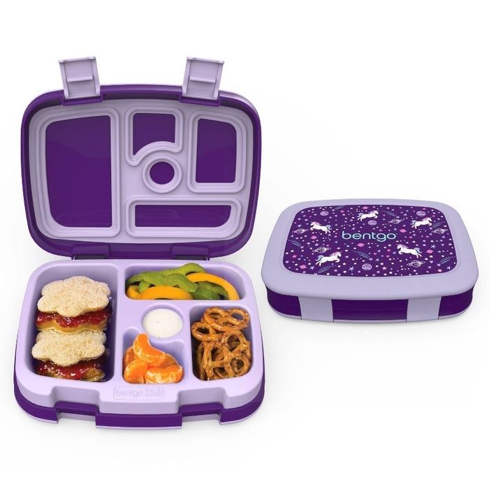 An open lunch box with compartments holding sandwiches, sliced peppers, dip, oranges, and pretzels; the purple lid features a unicorn design
