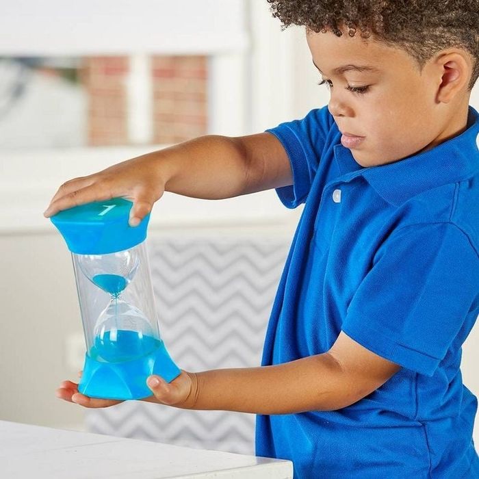 A child holds a large hourglass timer, observing the sand as it slowly falls. The object seems to be a toy or an educational tool.