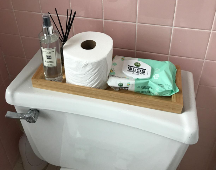 The reviewer's bamboo tray positioned on top of the toilet, holding toilet paper, wipes, and an oil diffuser
