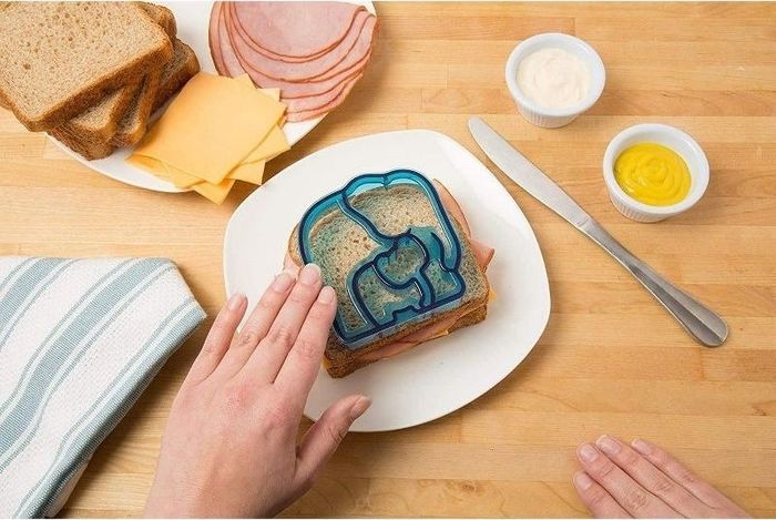A sandwich filled with deli meats and cheese is being shaped into a bear using a blue sandwich cutter. The bread, cheese, and condiments are spread out on a wooden surface.