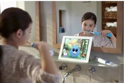 A young child brushes their teeth in front of a bathroom mirror while watching a tablet that shows a blue animated character demonstrating proper brushing techniques.