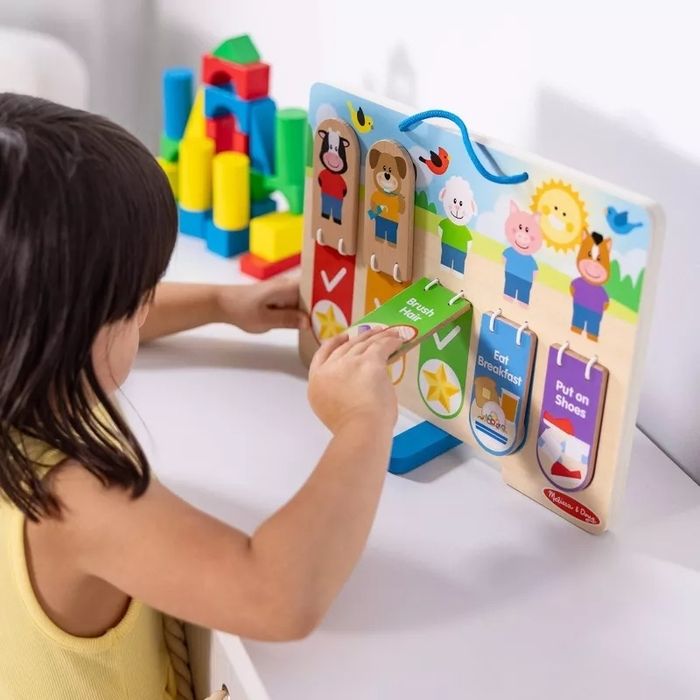 A young child in a yellow shirt interacts with a wooden chart depicting daily tasks, featuring animal characters and activities like brushing teeth and having breakfast