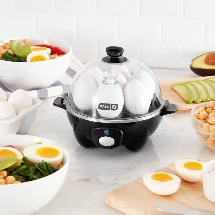 A Dash egg cooker rests on a kitchen counter, surrounded by bowls filled with fresh salads and chopped veggies, alongside some perfectly cooked hard-boiled eggs