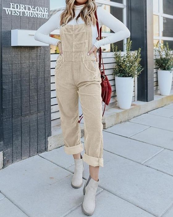A person wearing beige corduroy overalls, a white long-sleeve shirt, beige ankle boots, and carrying a red bag on a sidewalk outside a building.