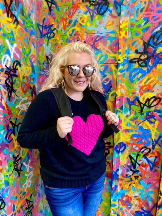 A reviewer wearing a black sweater with a pink heart design, ending just past the waist.