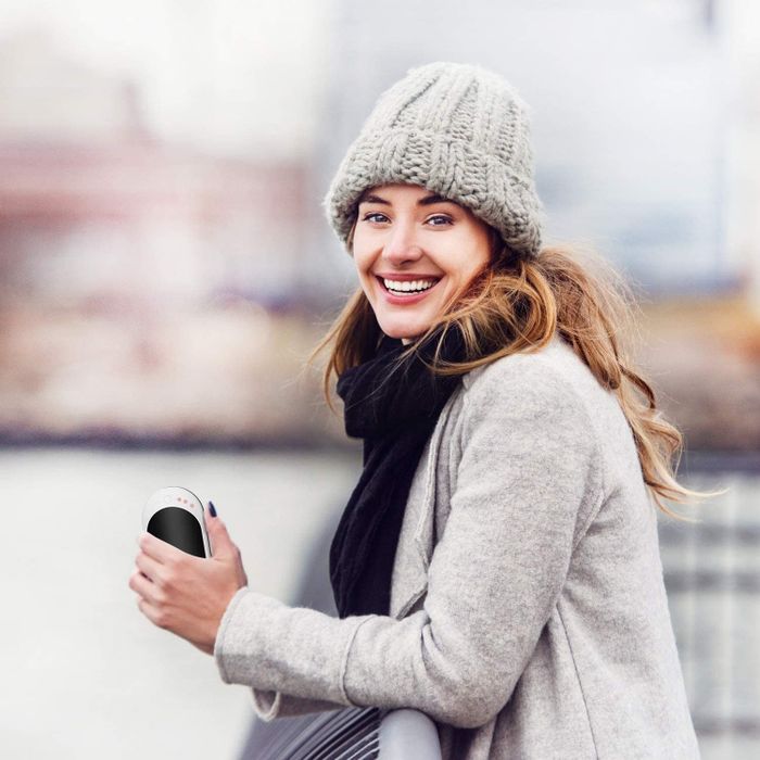 A model smiling while holding the Infray hand warmer in their left hand, standing outside on a cold day.