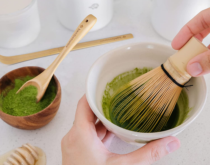 a hand using a bamboo whisk to mix matcha beside a bowl of loose matcha powder with a spoon resting inside