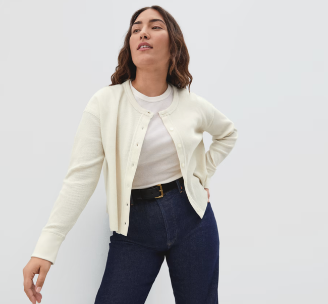 The model is wearing a soft cream cardigan layered over a white top and paired with denim jeans.