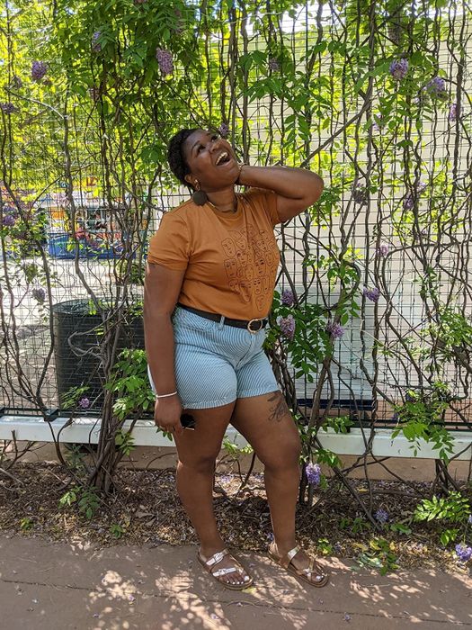 Reviewer is wearing a light orange top with abstract face designs paired with denim shorts