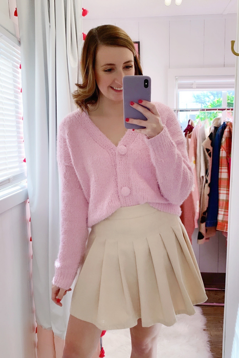 The model is seen wearing a soft pink fuzzy cardigan paired with a light cream pleated skirt.