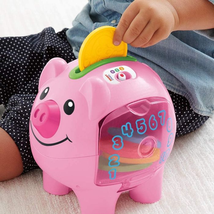 A toddler places a plastic coin into a cheerful pink piggy bank, learning about saving and counting in the process.