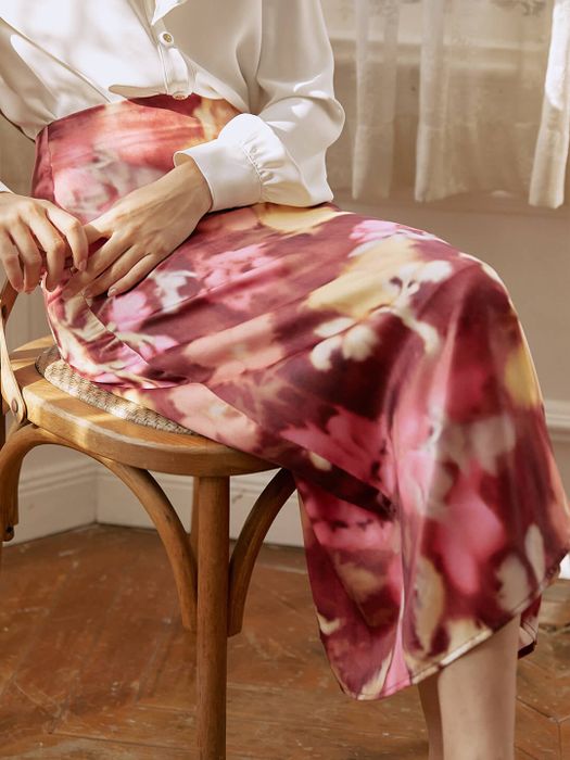 The model is dressed in a white top and a midi skirt featuring a vibrant red, pink, and yellow watercolor pattern.