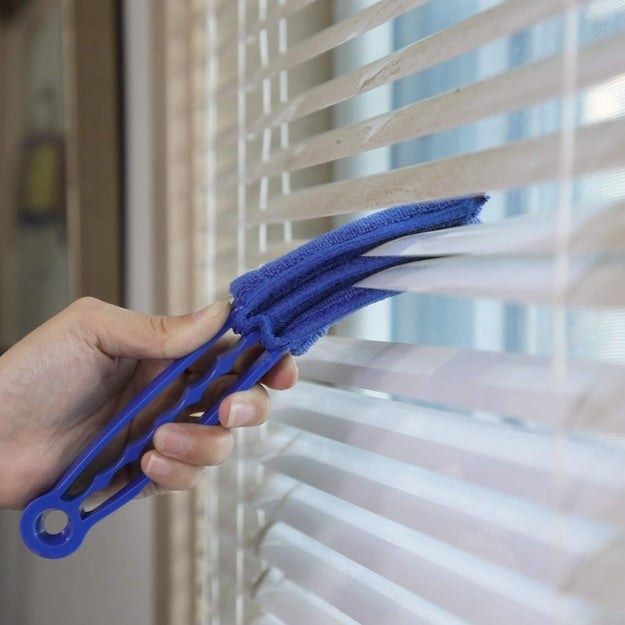 A hand grips a blue blinds duster, cleaning two window blinds simultaneously for efficient dusting.
