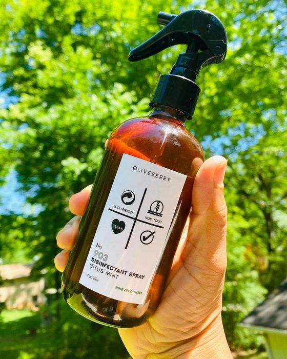 A hand holds a brown spray bottle labeled 'Oliveberry No. 902 Disinfectant Spray'.