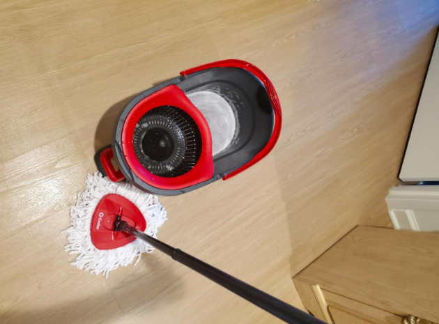 A reviewer uses a red and black microfiber spin mop to clean a hardwood floor.