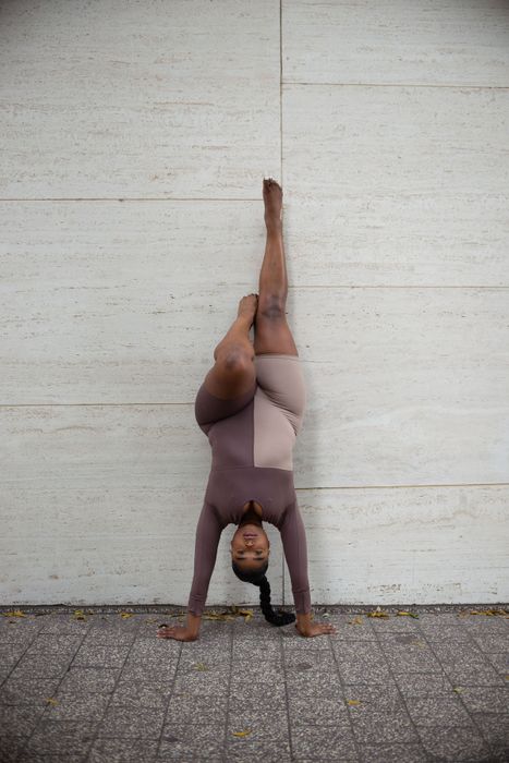 The model is wearing the two-tone long-sleeved bodysuit while performing a headstand against a wall.