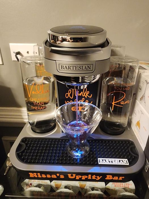 The Bartesian cocktail maker sits on a countertop with a glass next to it, surrounded by containers playfully labeled with quirky alcoholic phrases.
