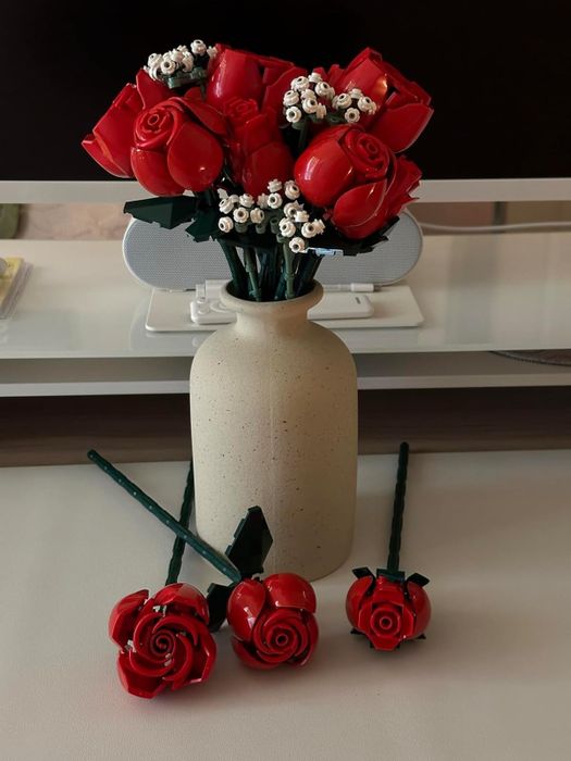 A ceramic vase displays a bouquet of LEGO roses along with baby's breath, while three additional LEGO rose stems rest on a nearby table