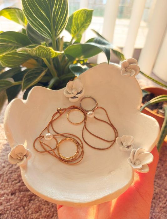 A white floral dish holding gold necklaces and rings with pearls, displayed in front of indoor plants, perfect for a shopping article