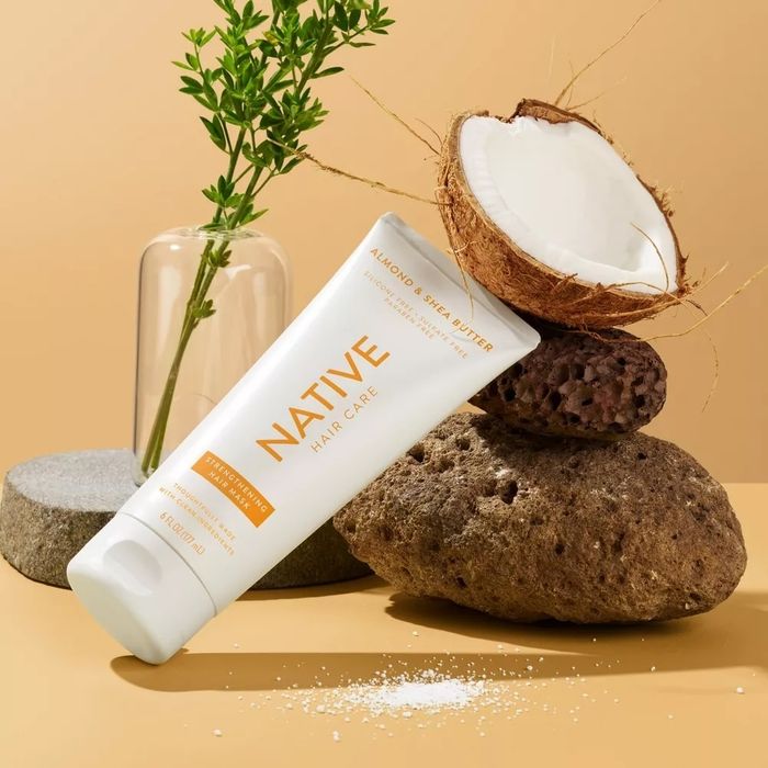 A tube of Native almond shea butter hair care placed on a rock, surrounded by a coconut half and lush greenery, highlighting the natural and nourishing qualities of the product.