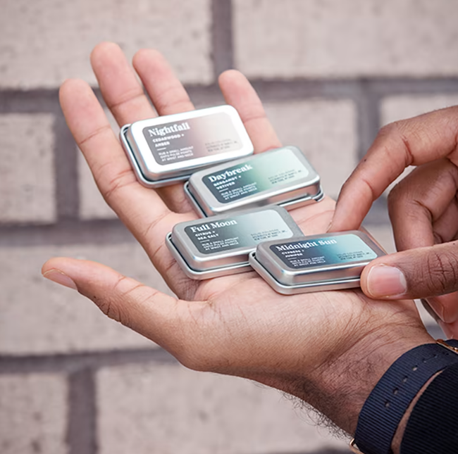 A hand holding four small tins labeled Nightfall, Daybreak, Full Moon, and Midnight Rain — most likely fragrances or skincare items
