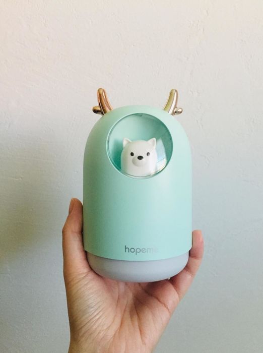 A hand holds the small humidifier, which is oval-shaped with tiny antlers on top and features a window showing a dog inside.