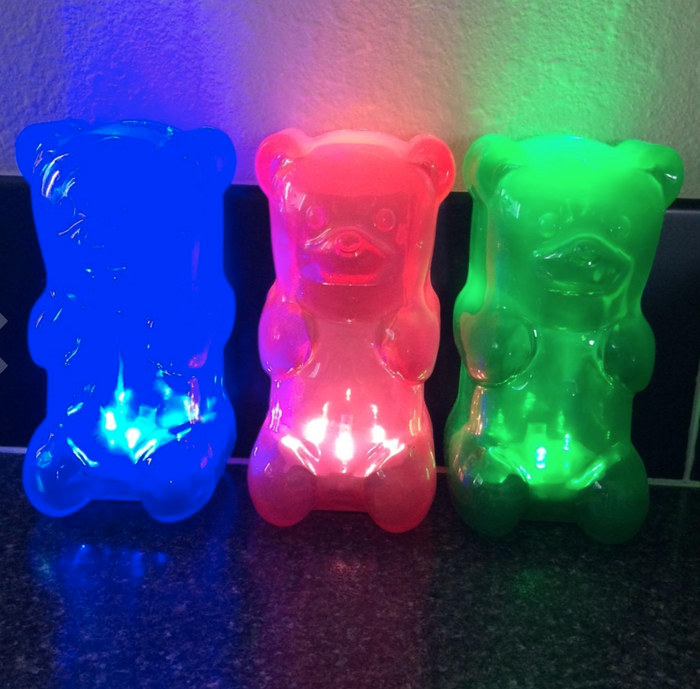 Three gummy bear-shaped night lights glowing in blue, red, and green, arranged side by side on a dark surface.