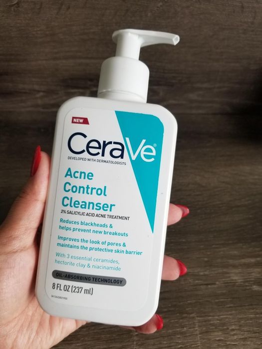 A reviewer holding an 8 fl oz bottle of CeraVe Acne Control Cleanser, containing 2% salicylic acid, designed to minimize blackheads and prevent new breakouts.