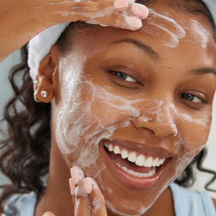 A person applies a foamy facial cleanser with both hands while smiling, wearing a headband and sporting multiple ear piercings.