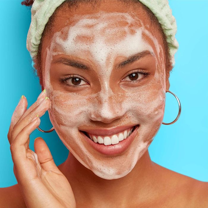 A model wearing a green headband, smiling while applying foamy facial cleanser.