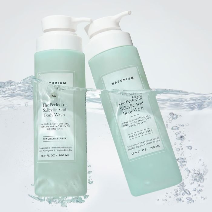 Two bottles of Naturium body wash, partially submerged in water, labeled as salicylic acid and fragrance-free, designed to smooth and soften the skin
